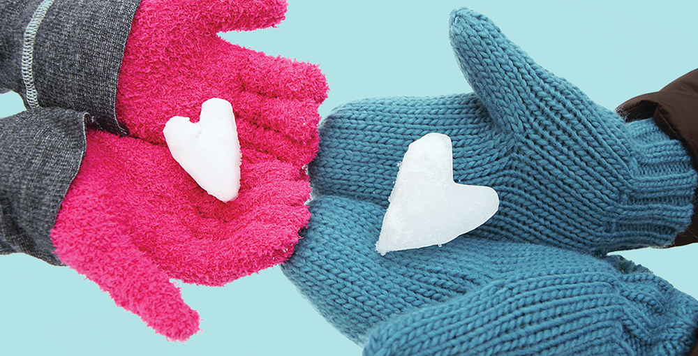 Two pairs of hands wearing winter gloves and mittens hold heart-shaped snow balls.