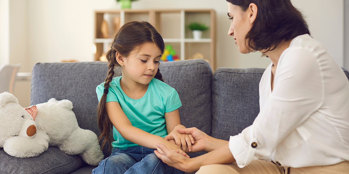 A mother and young daughter are holding hands as they talk sitting on a couch together