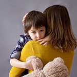 A mother hugs her son who is holding a teddy bear.